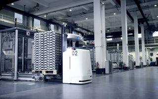 White high-lift automated guided vehicle beside a pallet stack in a plant aisle — ek robotics photo, illustrative