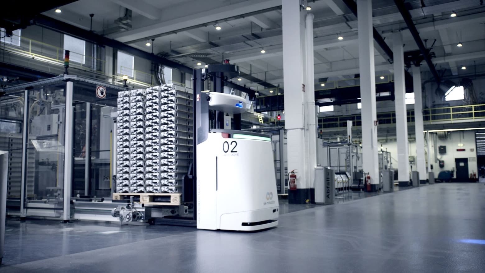 White high-lift automated guided vehicle beside a pallet stack in a plant aisle — ek robotics photo, illustrative