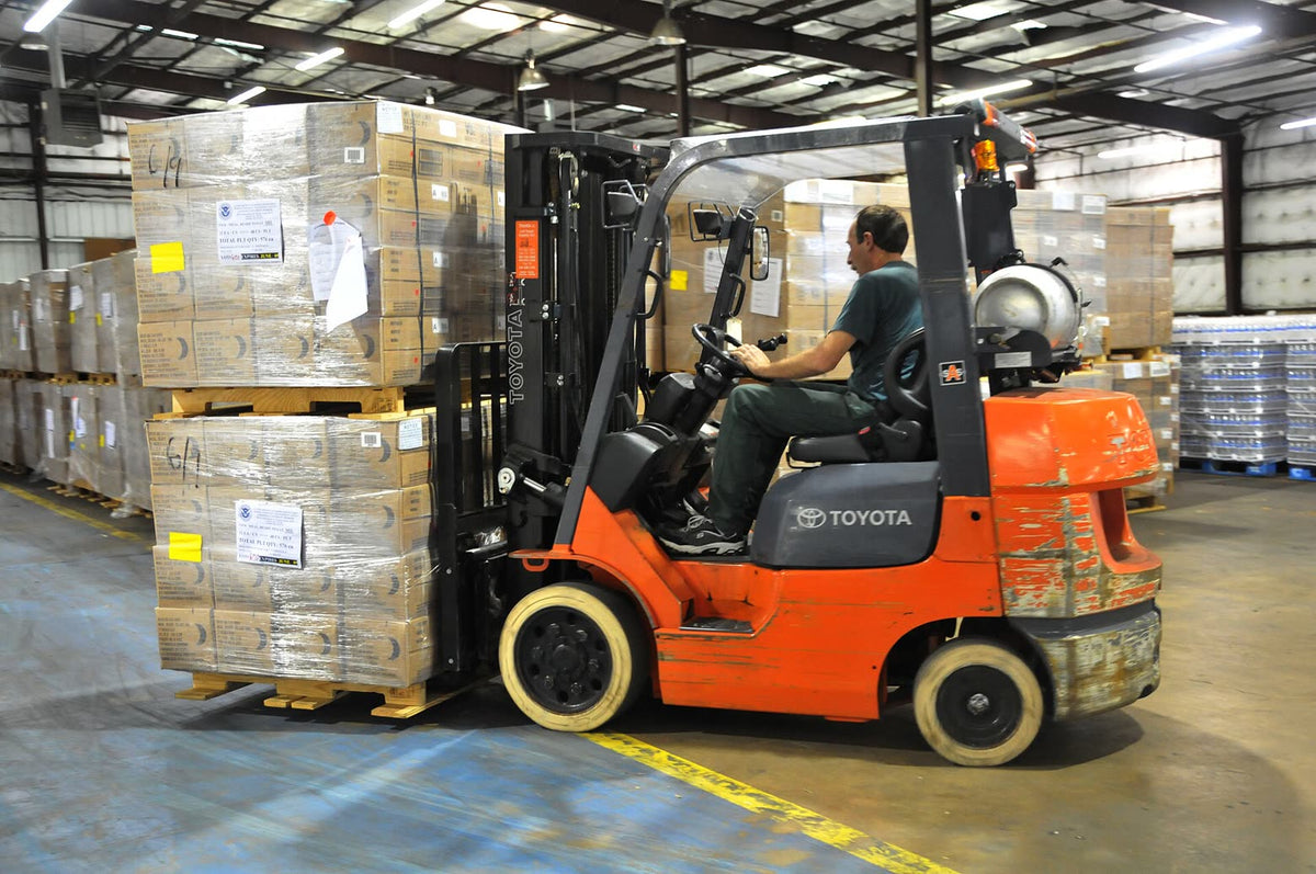 Operator driving a forklift with palletized cartons inside a warehouse — FEMA public-domain photo, illustrative